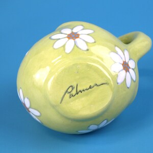 Puede incluir: Jarra de cer&aacute;mica verde claro, peque&ntilde;a y redonda, con asa. Margaritas blancas con centros naranjas pintadas alrededor de la jarra. La firma del artista, "Palmer", est&aacute; escrita en la base.