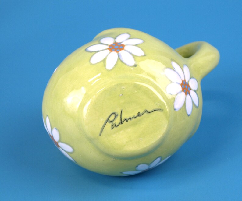 Puede incluir: Jarra de cer&aacute;mica verde claro, peque&ntilde;a y redonda, con asa. Margaritas blancas con centros naranjas pintadas alrededor de la jarra. La firma del artista, "Palmer", est&aacute; escrita en la base.