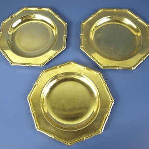 May include: Three gold-colored plates with an octagonal shape and bamboo-style edging. The plates have a shiny finish and are arranged on a blue surface. The plates are likely for decorative or serving purposes.