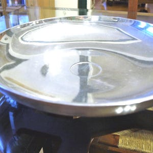 May include: A shiny, silver-colored serving tray with a shallow, rounded design. The tray reflects the surrounding environment, creating a mirrored effect. The tray is likely made of metal and is suitable for serving food or displaying items.