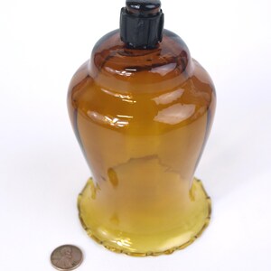 Puede incluir: Un portavelas de cristal &aacute;mbar transl&uacute;cido con una base ancha y festoneada y un cuerpo redondeado. Un tap&oacute;n de pl&aacute;stico negro est&aacute; insertado en la abertura superior. Una moneda de cobre est&aacute; colocada junto a la base como referencia de escala.