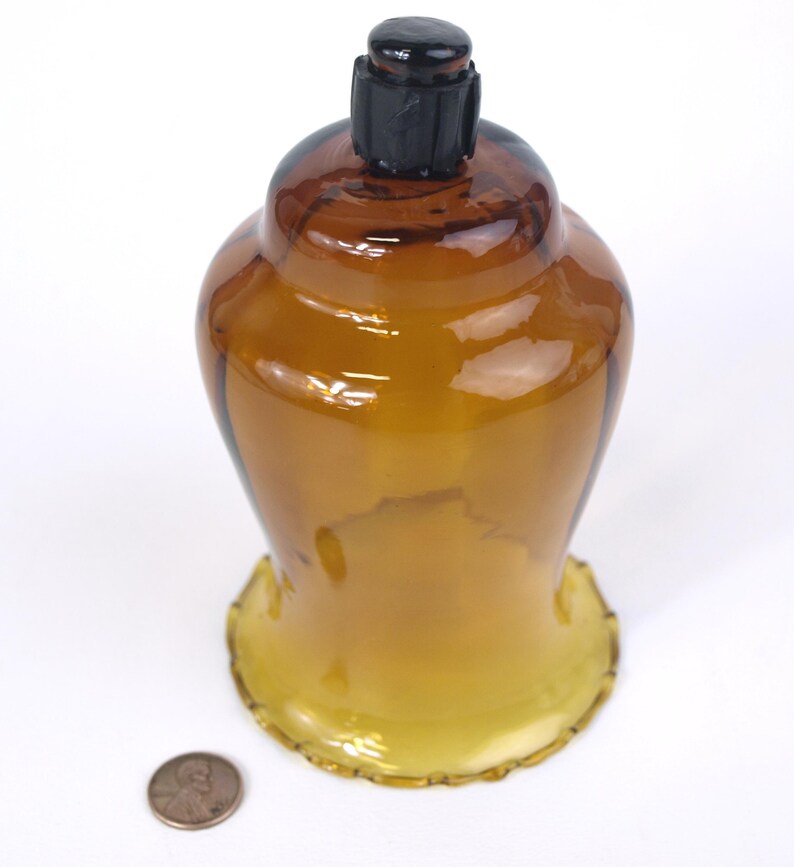 Puede incluir: Un portavelas de cristal &aacute;mbar transl&uacute;cido con una base ancha y festoneada y un cuerpo redondeado. Un tap&oacute;n de pl&aacute;stico negro est&aacute; insertado en la abertura superior. Una moneda de cobre est&aacute; colocada junto a la base como referencia de escala.