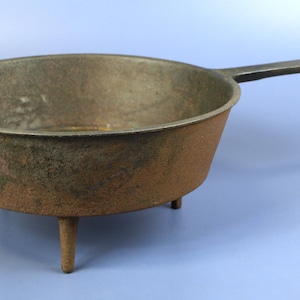 May include: A cast iron skillet with a long handle and three short legs. The pan has a round, shallow bowl shape with a dark, aged appearance. The handle has a hole at the end for hanging. The skillet is likely used for cooking.