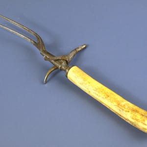 Op de afbeelding: Antieke vleesvork met een metalen vorkkop en een lichtgele handgreep. De handgreep heeft een cilindrische vorm en is afgedekt met een metalen uiteinde. De vork staat tegen een blauwe achtergrond.