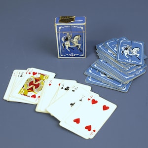 以下が含まれることがあります： 騎士が描かれた青と金の箱に入った、ヴィンテージのトランプセット。 デッキには、赤、黒、金色のスートのさまざまなトランプが含まれています。 カードは青い表面に配置されています。