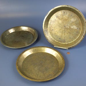 May include: Three vintage pie plates made of brass. The plates are different sizes and have a worn, aged look.