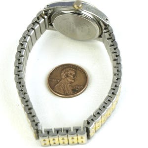 May include: A silver and gold-toned wristwatch with a stretch band, shown next to a penny. The watch has a round face with a silver bezel and a gold-colored crown. The band is a mix of silver and gold-colored links.