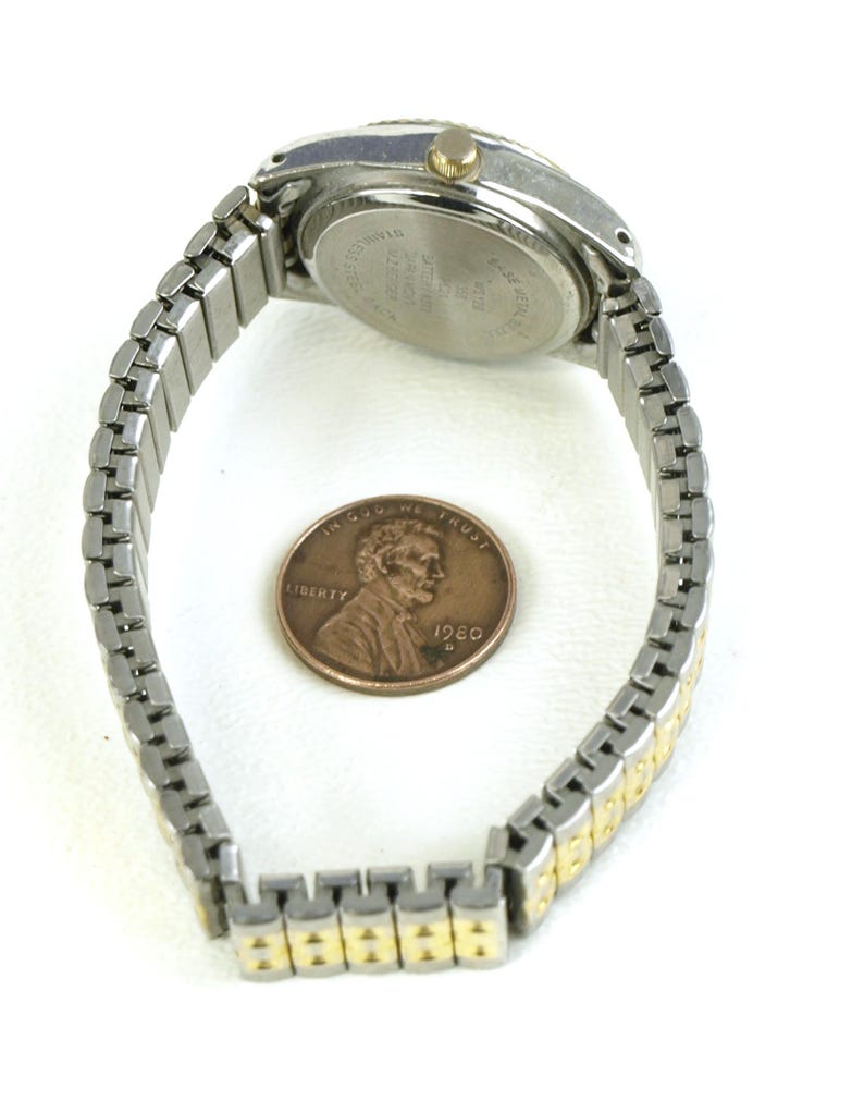 May include: A silver and gold-toned wristwatch with a stretch band, shown next to a penny. The watch has a round face with a silver bezel and a gold-colored crown. The band is a mix of silver and gold-colored links.