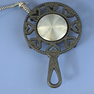 Puede incluir: Un salvamanteles circular de hierro fundido negro con un patrón decorativo de corazones recortados que rodea un disco central de metal plateado cepillado. Un cordón trenzado en blanco y negro con un resorte metálico plateado está unido arriba.