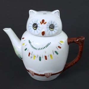 Może przedstawiać: Biały ceramiczny czajnik w kształcie sowy z malowanym wzorem w kolorze czerwonym, zielonym, żółtym i brązowym. Sowa ma duże czarne oczy z białymi i niebieskimi akcentami.
