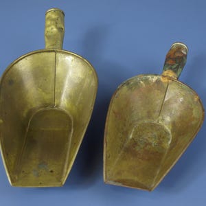 Peut inclure: Deux pelles anciennes en laiton avec des poignées, sur fond bleu. Les pelles ont une forme de bol arrondie et une poignée qui se courbe vers le haut. Le laiton a une teinte dorée chaude avec des zones de patine.