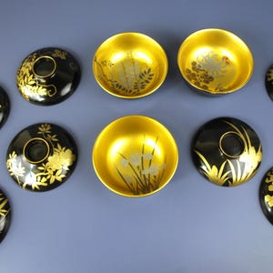 May include: A collection of black and gold lacquered bowls and lids. The lids display detailed gold floral and bamboo patterns. The bowls are gold-coloured with subtle floral designs. The items are arranged on a blue surface.