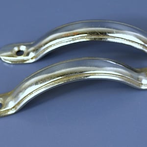 Könnte beinhalten: Zwei silberfarbene, gebogene Schrankgriffe. Jeder Griff hat ein abgerundetes, gewölbtes Design mit einem Loch an jedem Ende zur Befestigung. Die Griffe sind vor einem gedämpften blauen Hintergrund angeordnet.