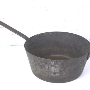 May include: A dark gray, antique-style metal pan with a long handle and a small loop handle on the side. The pan has a wide, open bowl shape and appears to be made of heavy-duty metal. The pan is likely used for cooking.