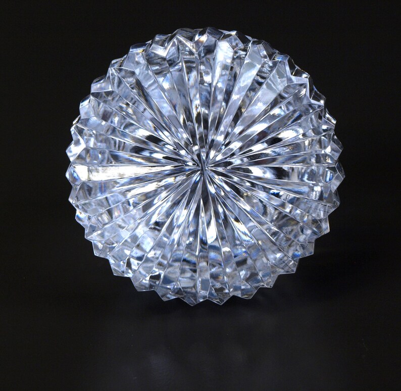 May include: A clear, faceted glass object with a radial design. The object is round and appears to be a decorative piece, possibly a paperweight or a decorative bowl. The facets create a play of light and shadow.