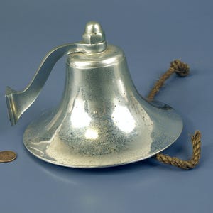 May include: A silver-colored bell with a curved handle and a rope attached. The bell has a wide base and a rounded top. A penny is next to the bell for size comparison. The bell is likely made of metal.