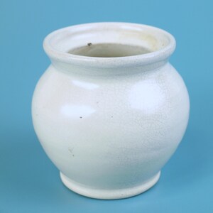 May include: A small, off-white ceramic pot with a rounded body and a slightly flared rim. The pot has a crackled glaze, giving it a vintage appearance. The pot sits on a small, circular base. The background is a solid light blue.