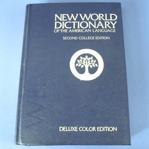 May include: A dark blue hardcover book titled New World Dictionary of the American Language, Second College Edition, Deluxe Color Edition. The cover features a white circular emblem containing a stylized tree graphic centered on the front.