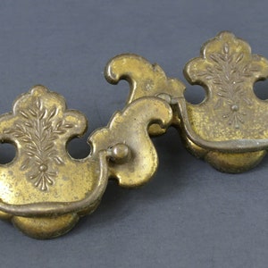 Puede incluir: Dos tiradores de armario de latón antiguo con diseños florales ornamentados y un asa curva. Los tiradores tienen un acabado desgastado y envejecido.