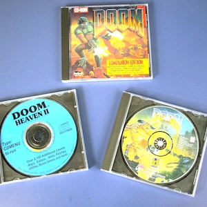 May include: Three CD-ROMs of the classic video game "Doom." The top case displays "Doom Companion Edition" with a green-armored character. The other two cases show the game discs, one labeled "Doom Heaven II."