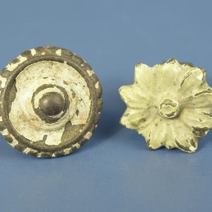 May include: Two decorative cabinet knobs. One is round with a central knob and a distressed white and brown finish. The other is a flower shape with a similar distressed finish. Both are on a blue background.