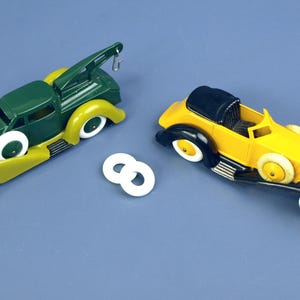 Puede incluir: Dos coches de juguete antiguos, un camión de remolque verde y amarillo y un descapotable amarillo y negro, están sobre una superficie azul. Ambos tienen ruedas blancas. Dos anillos blancos están entre los coches.