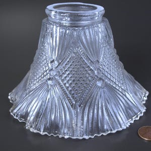 May include: Clear glass lampshade with a diamond and fan pattern. The lampshade has a flared shape with a ruffled edge. A penny is next to the lampshade for size comparison.