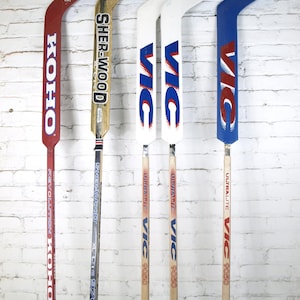 May include: Five vintage hockey sticks. The sticks have red, white, blue, and gold blades with various brand names, including KOHO, SHER-WOOD, and VIC. The sticks are displayed against a white brick wall.