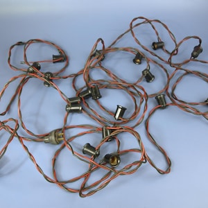 Puede incluir: Una enredada cadena de luces navideñas vintage con cables trenzados rojos y verdes. Las luces tienen pequeños casquillos de metal oscuro y están dispersas sobre una superficie azul claro. El cable tiene un enchufe macho.