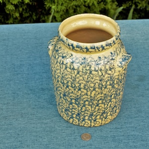 Puede incluir: Un tarro de cerámica amarillo con motas azules y una boca ancha. El tarro tiene dos asas a los lados y está sentado sobre una superficie azul.