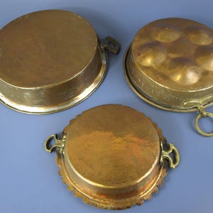 May include: Three antique copper cooking pans. The pans are round with a hammered texture and brass handles. One pan has a domed lid with circular indentations. The pans are displayed on a blue surface.