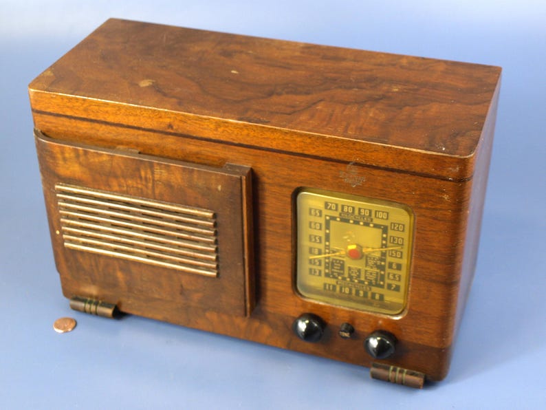 Puede incluir: Una radio vintage de madera con un dial dorado y botones negros. La radio tiene una rejilla de altavoz en el lado izquierdo y un gabinete de madera.