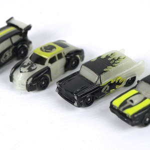 Puede incluir: Cuatro coches de juguete en miniatura con pintura que brilla en la oscuridad. Los coches son negros con detalles amarillos y gráficos de calaveras.