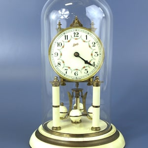 May include: A Schatz torsion clock featuring a cream-colored face with black numerals and a floral garland design, encased in a clear glass dome. The clock has a brass frame, cream-colored pillars, and a rotating pendulum with four white spheres.