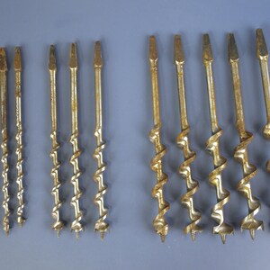 May include: A collection of antique auger drill bits, varying in size and design. The bits have a spiral shape with a pointed tip and are made of a metallic material with a weathered, aged appearance. A penny is included for size reference.