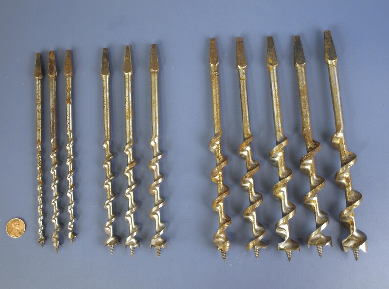May include: A collection of antique auger drill bits, varying in size and design. The bits have a spiral shape with a pointed tip and are made of a metallic material with a weathered, aged appearance. A penny is included for size reference.