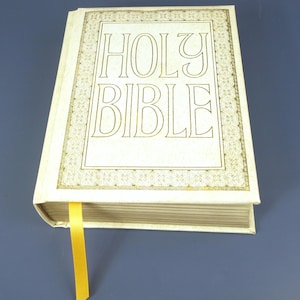 Könnte beinhalten: Eine cremefarbene Heilige Bibel mit goldfarbenen Buchstaben und einem dekorativen Rand. Die Worte "HOLY BIBLE" sind auf dem Einband zu sehen. Ein gelbes Leseband ragt aus den Buchseiten heraus.