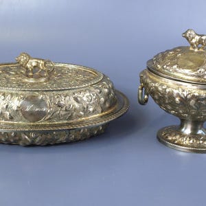 May include: Two antique silver serving dishes. One is oval with a lion finial and floral detailing. The other is a lidded bowl with a lion finial, ornate handles, and a pedestal base. Both are on a gray surface.