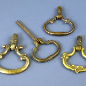 May include: Four antique brass drawer pulls with ornate designs. The pulls have a curved handle shape and are in various states of attachment. The brass has a warm, aged patina, suggesting vintage or salvaged hardware.