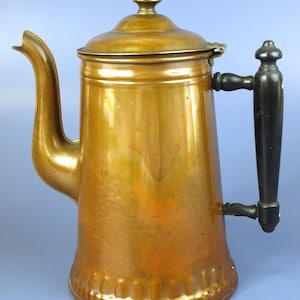 Puede incluir: Una cafetera de cobre con un asa de madera negra. La cafetera tiene una tapa con un pequeño adorno y un pico curvo. El cobre presenta una pátina cálida y envejecida. El diseño es clásico y funcional. Altura aproximada de 25 cm.