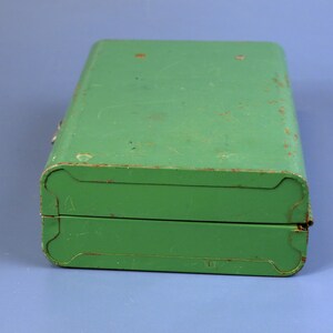 May include: A vintage, rectangular green metal box with rounded corners and a hinged lid. The box shows signs of wear, with visible scratches and rust, suggesting age and use. The box is closed and sits on a blue surface.