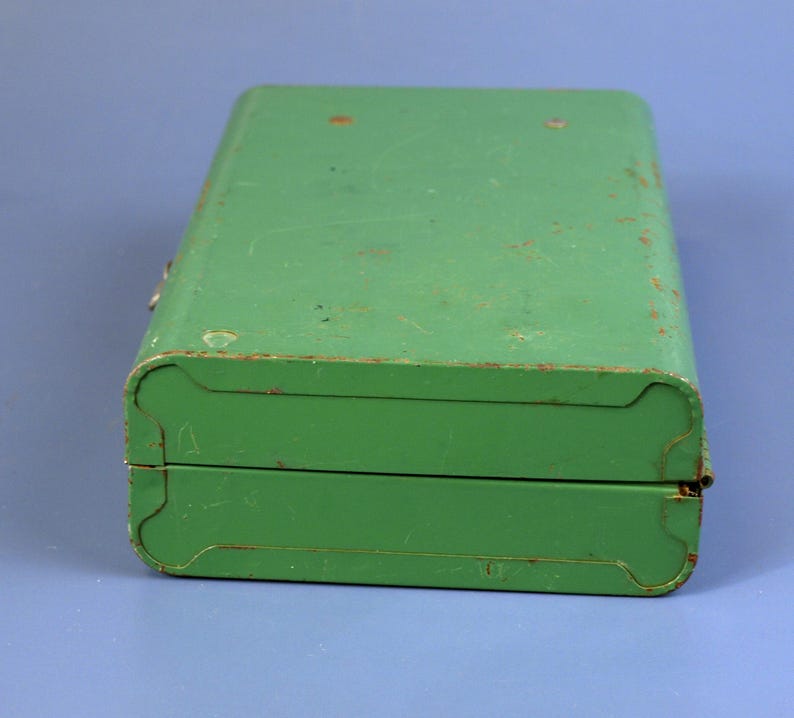 May include: A vintage, rectangular green metal box with rounded corners and a hinged lid. The box shows signs of wear, with visible scratches and rust, suggesting age and use. The box is closed and sits on a blue surface.