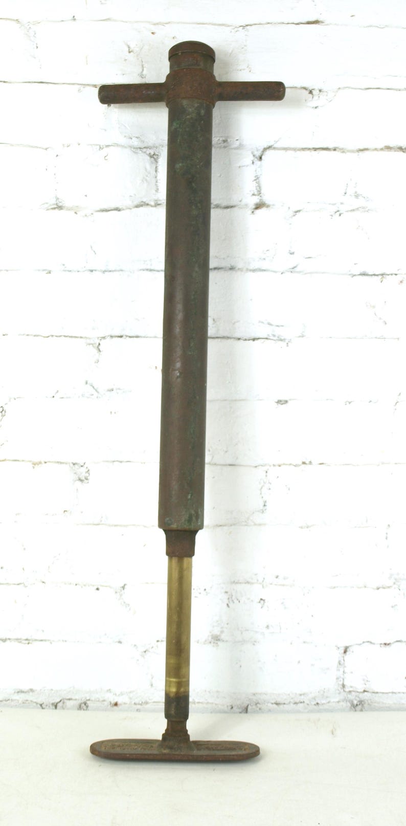 May include: A vintage manual hand pump featuring a long, weathered dark metal cylindrical body with a T-shaped handle at the top. The lower section consists of a polished brass rod extending down to a flat, oval-shaped metal base plate.