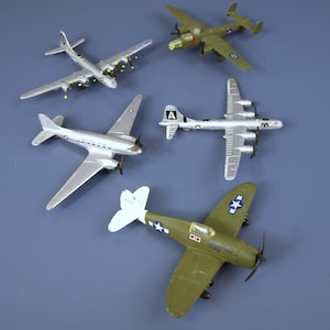 May include: A collection of toy airplanes in various designs and colors, including silver, olive green, and gray. The planes feature propellers, wings, and detailed markings, set against a blue background. The collection includes a variety of aircraft.
