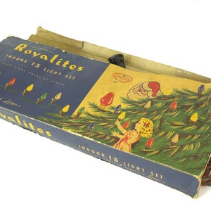 Könnte beinhalten: Vintage-Verpackung für Royalites Indoor 15 Light Set Weihnachtslichter. Die Schachtel zeigt eine farbenfrohe Illustration eines Weihnachtsbaums mit Lichtern und einem Kind, das ihn schmückt. Die Schachtel ist blau und weiß mit dem Markennamen und der Produktbeschreibung bedruckt.