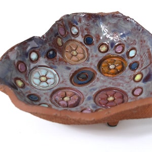 May include: A handmade ceramic bowl with a rustic brown exterior and a purple-grey interior. The bowl is decorated with colorful flowers and circles.