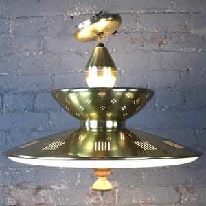 May include: A brass pendant light fixture featuring a tiered design with a perforated bowl section, a wide lower shade with vertical slit cutouts, and a white diffuser panel at the base. The fixture includes a central wooden finial at the bottom.