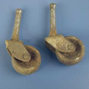 Puede incluir: Dos ruedas metálicas antiguas con aspecto oxidado y envejecido. Cada rueda tiene una rueda pequeña, un soporte giratorio y un vástago cilíndrico. Las ruedas parecen tener unos 3,8 cm de diámetro, lo que sugiere que son para un mueble pequeño.