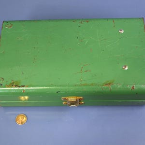 May include: A rectangular, vintage green metal box with a gold-colored clasp. The box shows signs of wear, with scratches and rust spots. A penny is next to the box for size comparison.