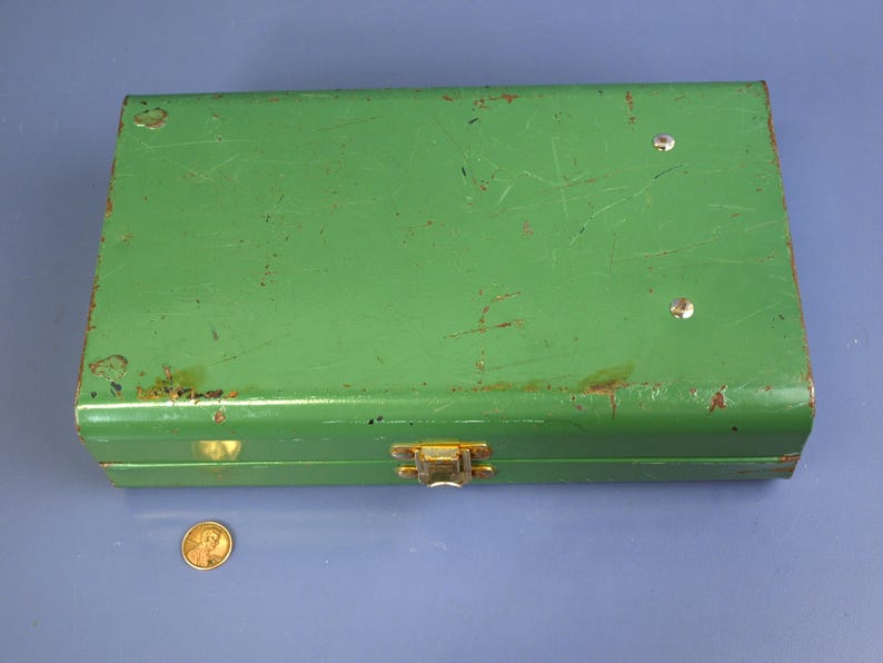 May include: A rectangular, vintage green metal box with a gold-colored clasp. The box shows signs of wear, with scratches and rust spots. A penny is next to the box for size comparison.